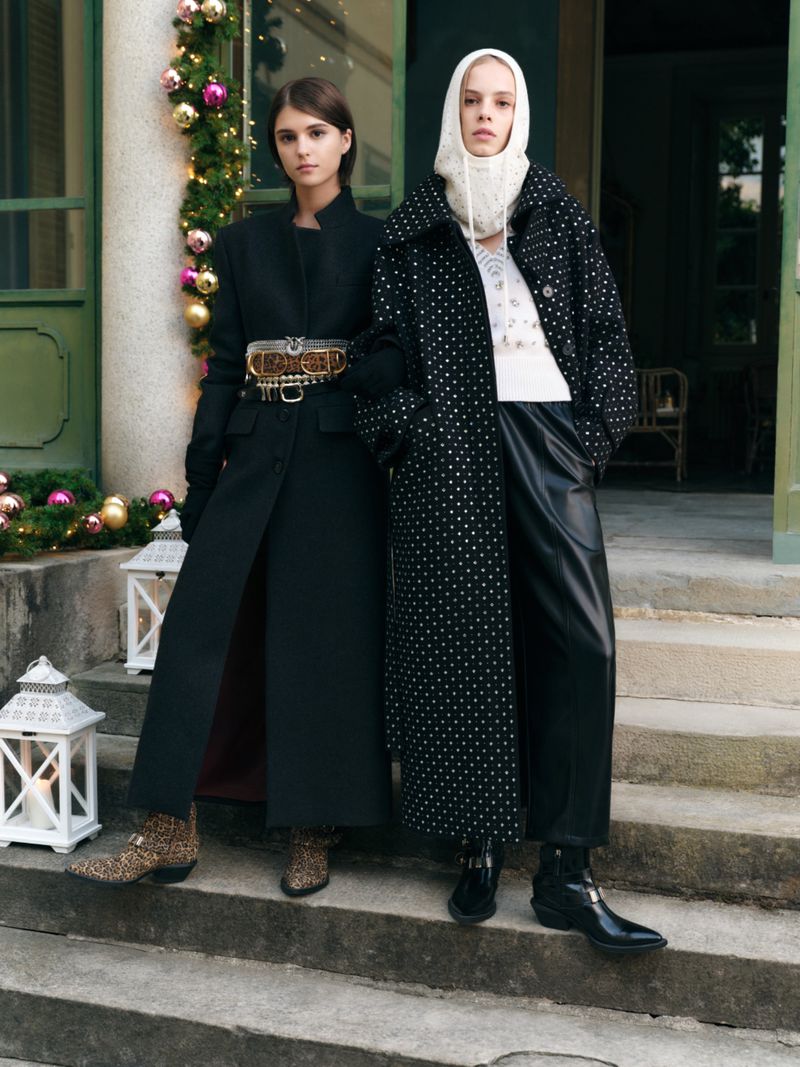 Two women pose on stone steps in long coats with Christmas decor in the background, showcasing a fashion editorial style and winter apparel.