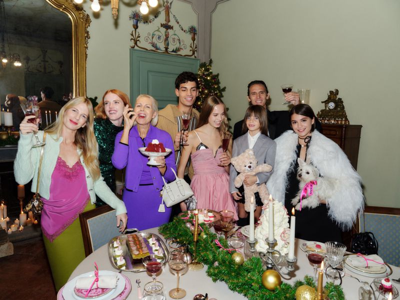 Group gathers indoors at a decorated table with food, drinks, and presents, celebrating with diverse ages and elegant attire.