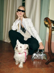 Estelle Veron in a white embellished blazer and black sunglasses crouching with a small white terrier and a metallic silver handbag.