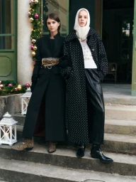 Two female models posing on stone steps outdoors wearing dark coats and boots, flanked by holiday decorations and lanterns.
