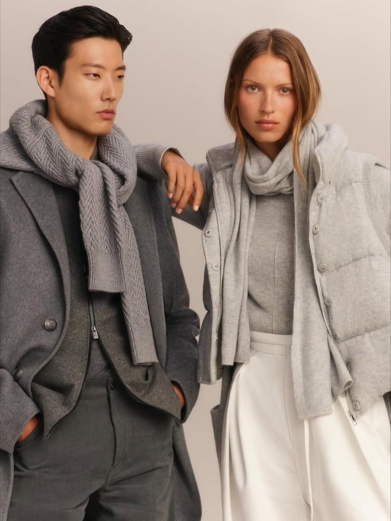 Two fashion models showcase winter apparel in a studio: a man in a grey coat and scarf, and a woman in a grey vest and scarf.