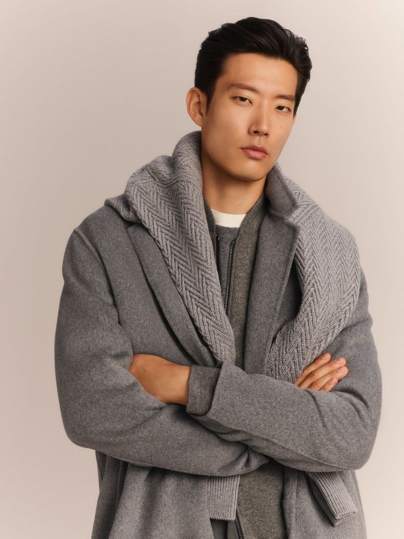 Portrait of a young adult male in a grey coat and scarf, posing indoors against a neutral backdrop in a fashion setting.