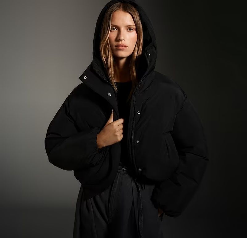 A woman in a black hooded puffer jacket looks directly at the camera against a dark grey gradient backdrop in a studio shot.