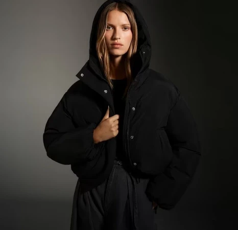 A woman in a black hooded puffer jacket looks directly at the camera against a dark grey gradient backdrop in a studio shot.