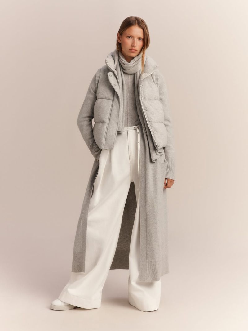 Fashion shot of a woman in a grey puffer vest, long grey coat, turtleneck, and white pants in a neutral indoor setting with soft lighting.