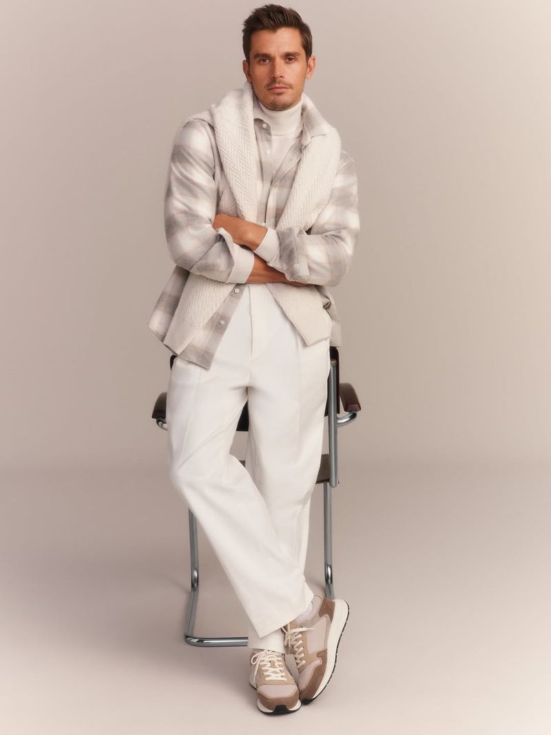 Full-length shot of an adult male leaning against a chair, wearing a beige turtleneck, patterned shirt, white trousers, and sneakers.
