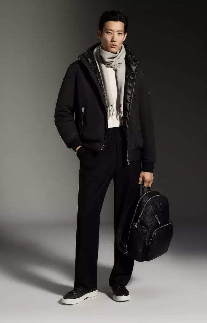 A full-length portrait of a male model wearing a black hooded jacket, scarf, trousers, and sneakers while holding a black backpack.