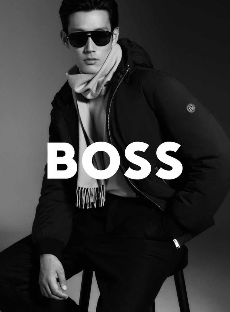 Asian male model in black puffer jacket, pants, beige scarf and sunglasses poses in studio with BOSS logo. Fashion photography.