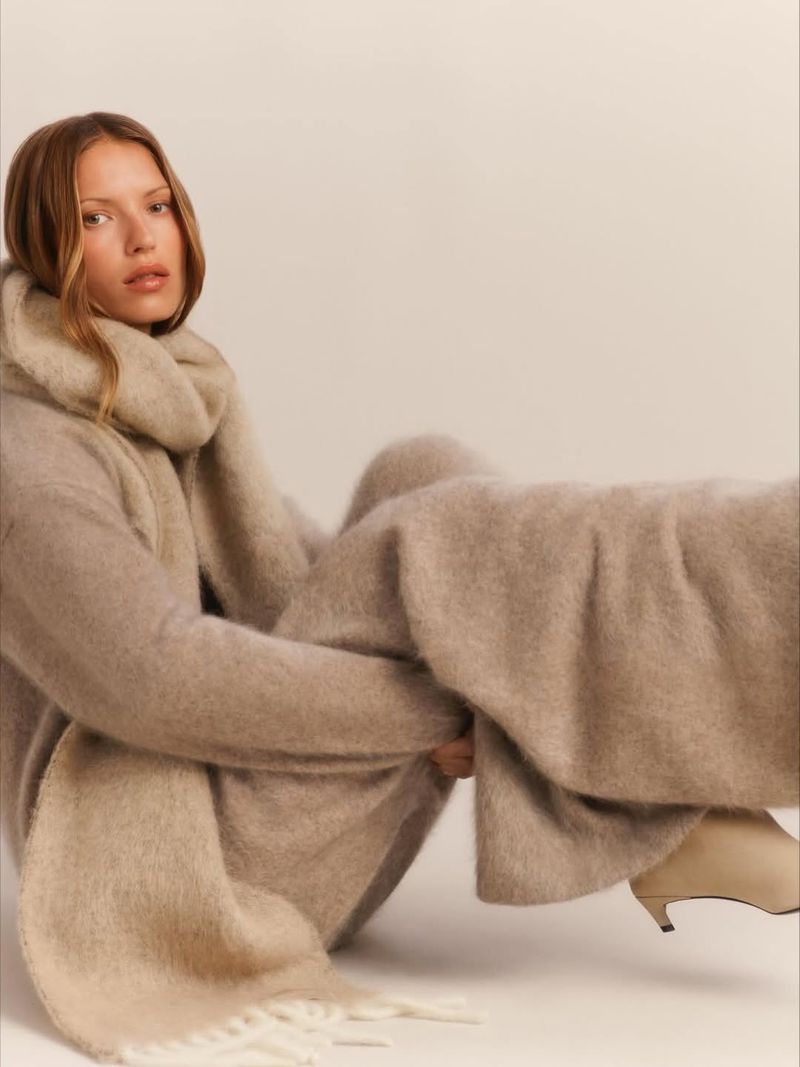 Fashionable adult woman in a sweater and trousers sits with a beige scarf indoors, showcasing a minimalist and stylish aesthetic.