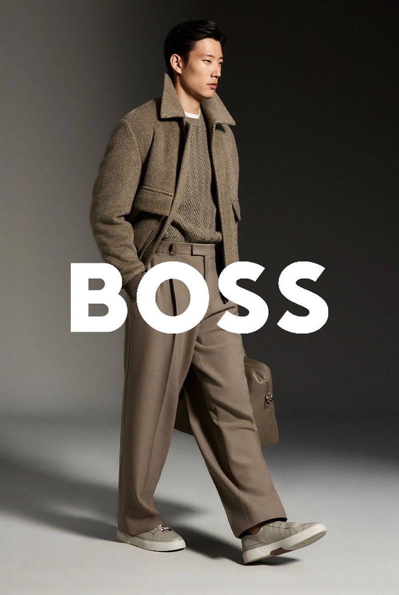 Asian male fashion model in a brown jacket, sweater, and pants holding a bag in a studio setting with the BOSS logo overlayed.