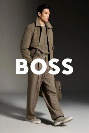 Asian male fashion model in a brown jacket, sweater, and pants holding a bag in a studio setting with the BOSS logo overlayed.