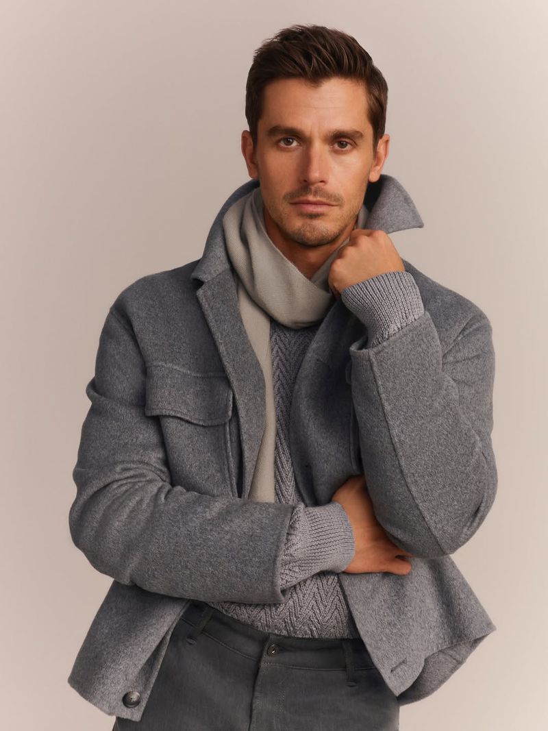 Close-up shot of a stylish man wearing a grey wool jacket over a knitted sweater and a light-colored scarf, posing in a photo studio.