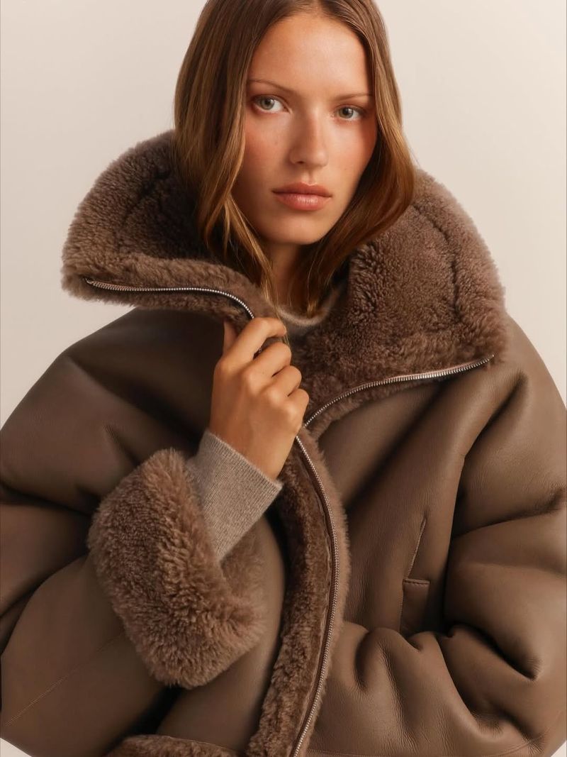 Fashion shot of a woman wearing a brown, oversized, sherpa-lined aviator jacket looking directly at the camera in an indoor setting.