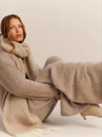 Model seated on the floor wearing a beige knit sweater, matching scarf and trousers against a neutral background.