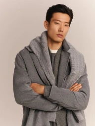Man wearing a grey coat with a knitted scarf draped over his shoulders against a neutral grey studio background.