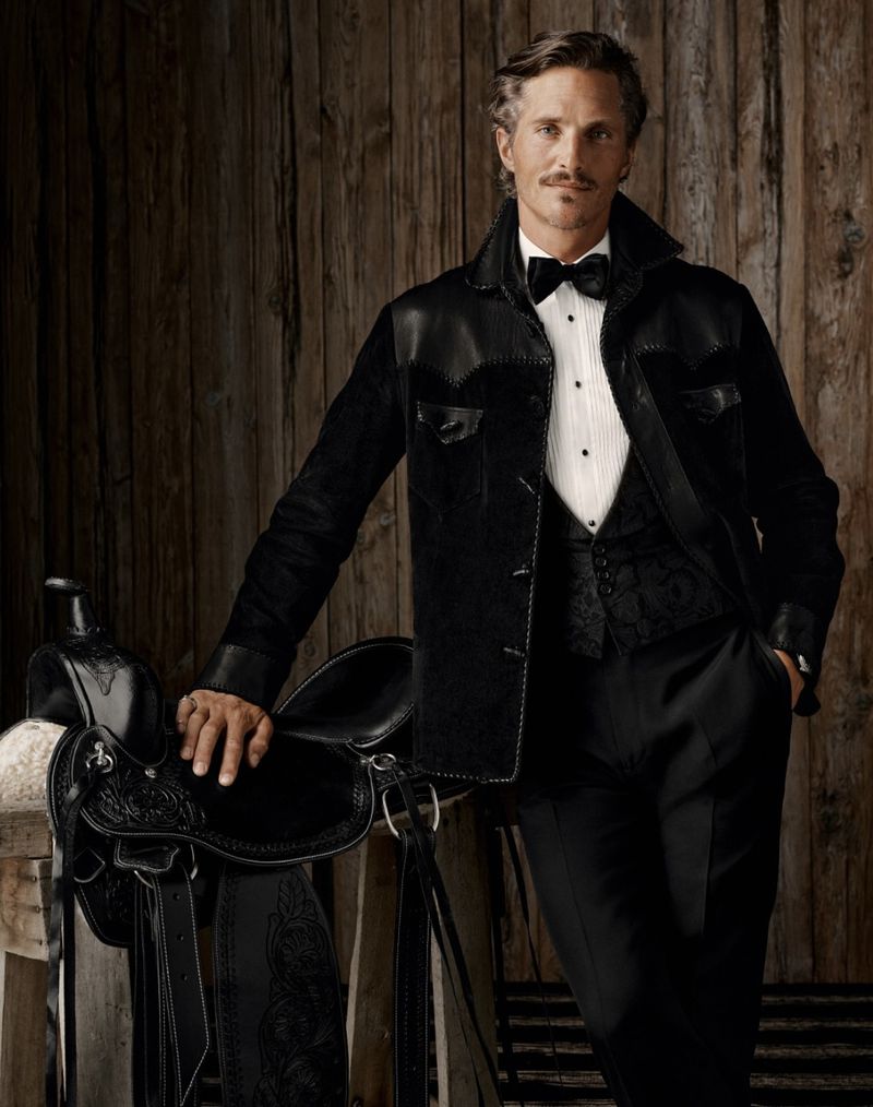 Adult male in formal wear with a bow tie poses with a saddle in front of a wooden wall in a studio, showcasing fashion and style.