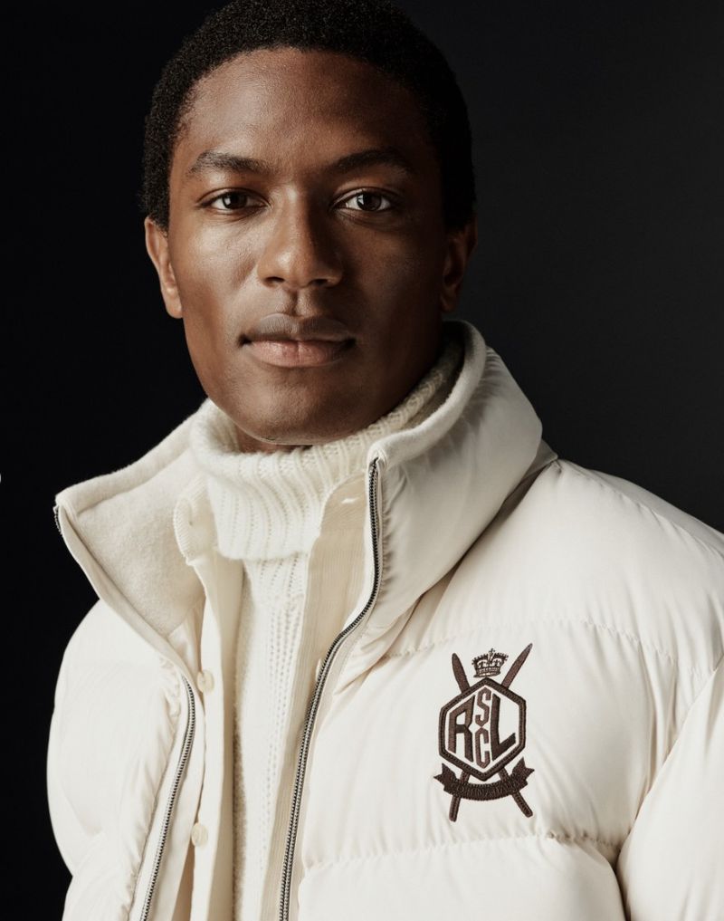 Headshot of a dark-skinned man wearing a cream-colored puffer jacket and turtleneck sweater against a dark background. Fashion shot.