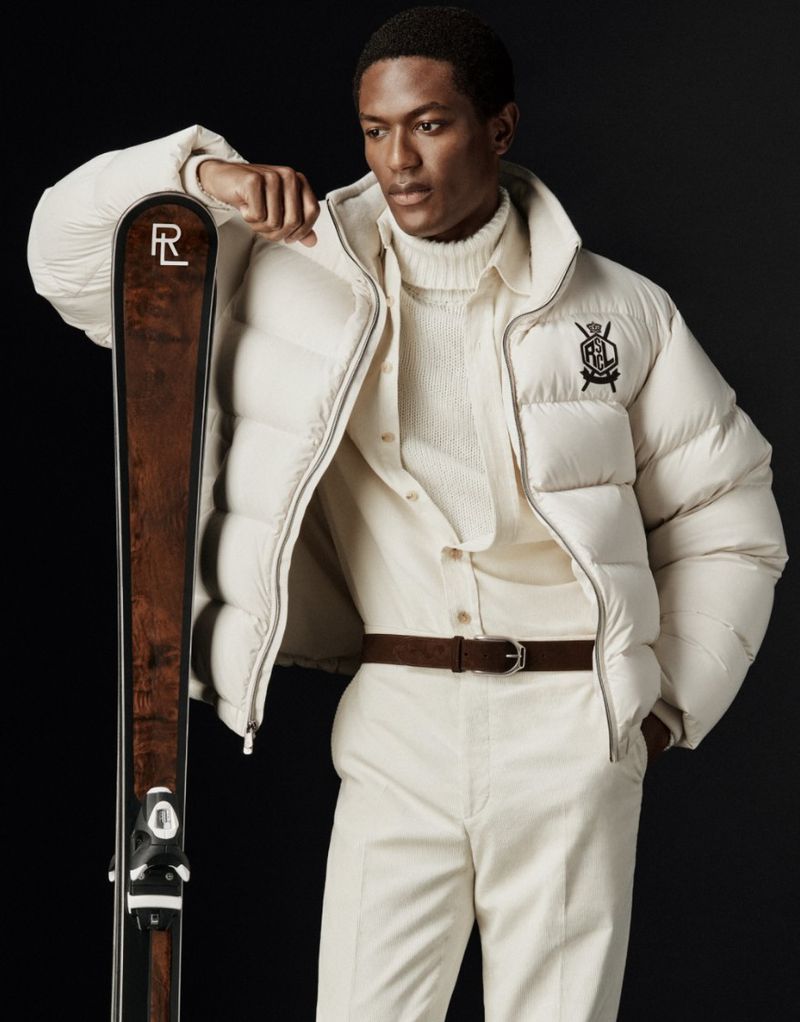 African American male in off-white puffer jacket leaning on a ski. He wears matching pants, shirt and turtleneck. Black backdrop.