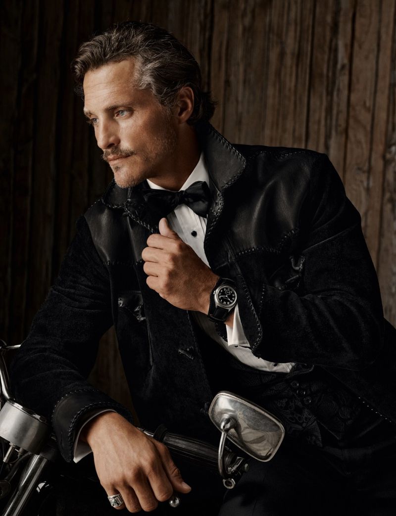 Portrait of a mature adult male with a moustache, wearing a black suit with a bowtie, posing on a black motorcycle.