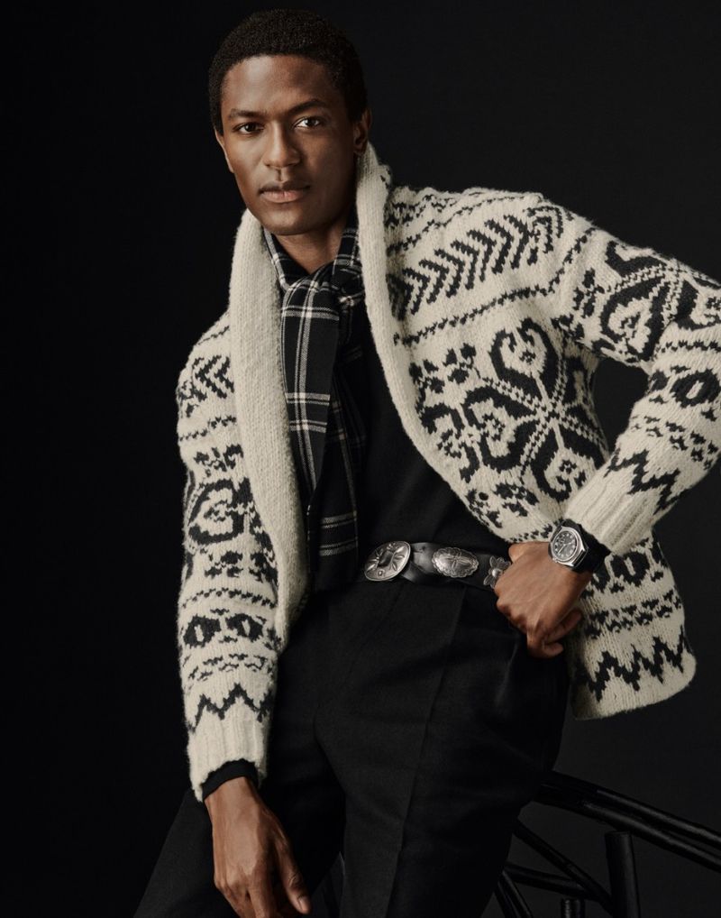 A handsome Black male model poses in a patterned cardigan, black shirt, and trousers against a dark backdrop for a fashion editorial.