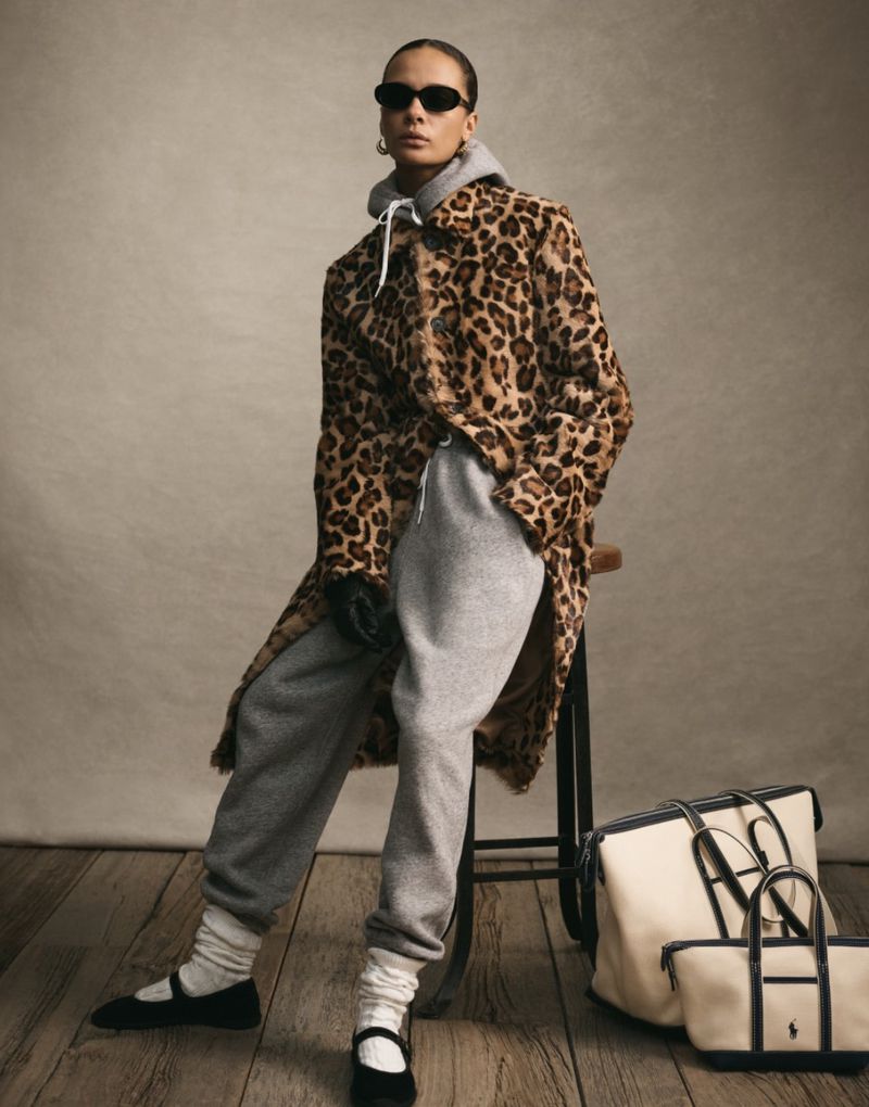 Woman in a leopard print coat, gray sweatsuit, and sunglasses poses in front of a neutral backdrop with two tote bags.