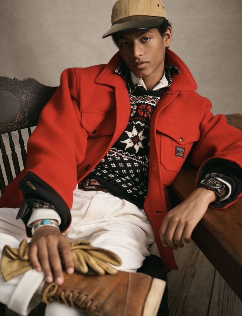 Young man seated wearing a red jacket, patterned sweater, and cap, accessorized with gloves and boots, exudes stylish casualness.