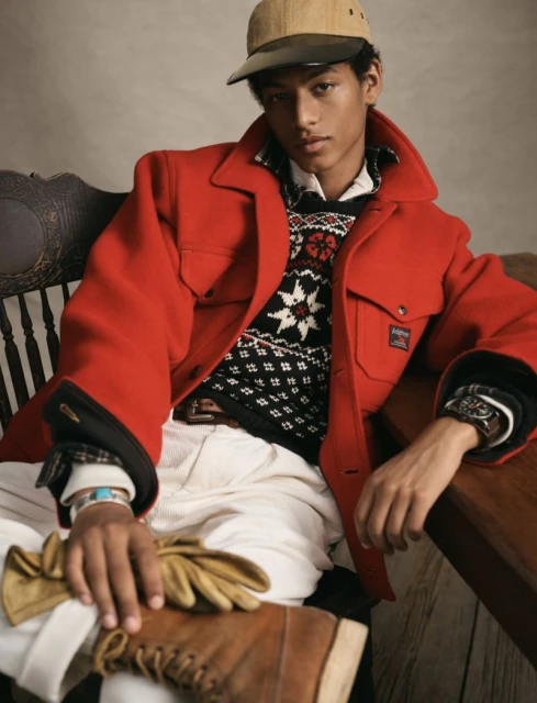 Young man seated wearing a red jacket, patterned sweater, and cap, accessorized with gloves and boots, exudes stylish casualness.