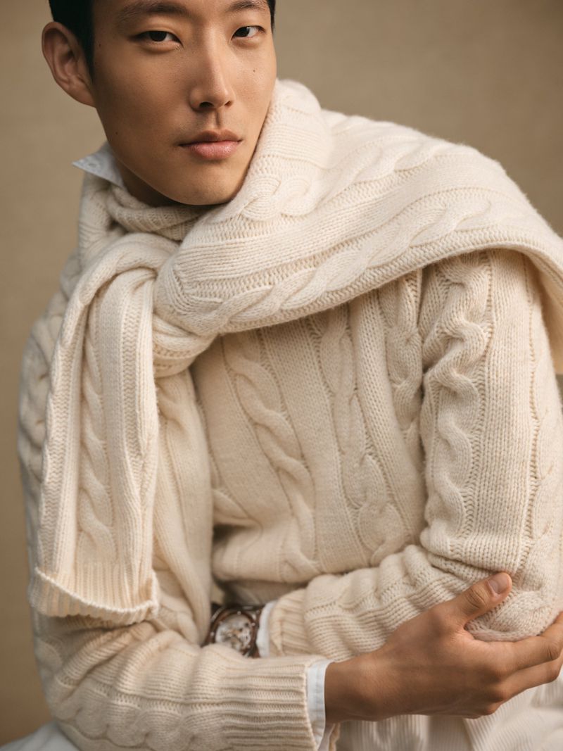 Portrait of an Asian male model wearing a cream-colored cable-knit sweater and scarf, posing in a softly lit studio setting.