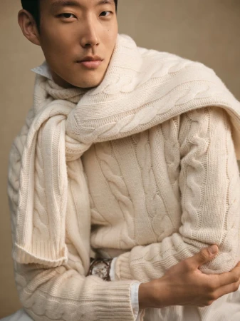 Portrait of an Asian male model wearing a cream-colored cable-knit sweater and scarf, posing in a softly lit studio setting.