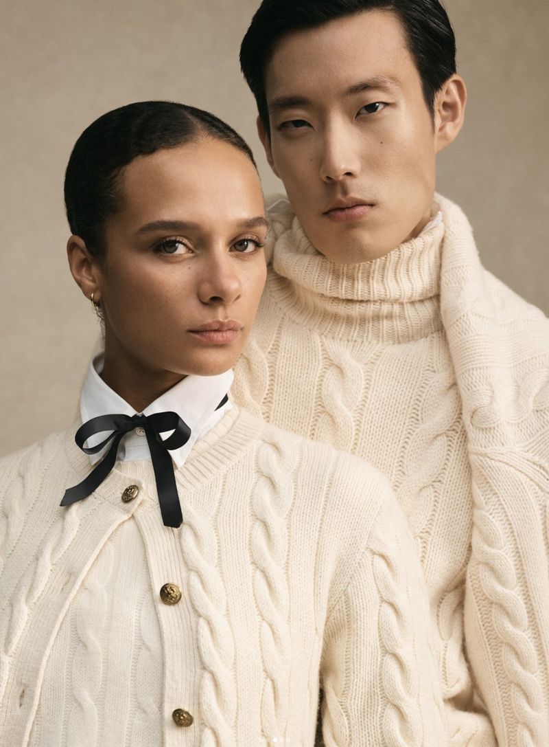 Fashion shot of a woman in a cream-colored cable knit cardigan with a black ribbon and a man in a turtleneck sweater.