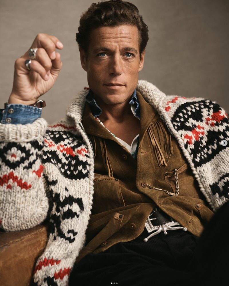 Portrait of a handsome adult man wearing a patterned cardigan sweater and suede jacket, posing indoors against a neutral backdrop.