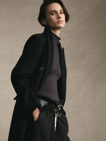 Model with short dark hair wearing a black tailored coat over a high-neck grey top against a neutral studio backdrop.