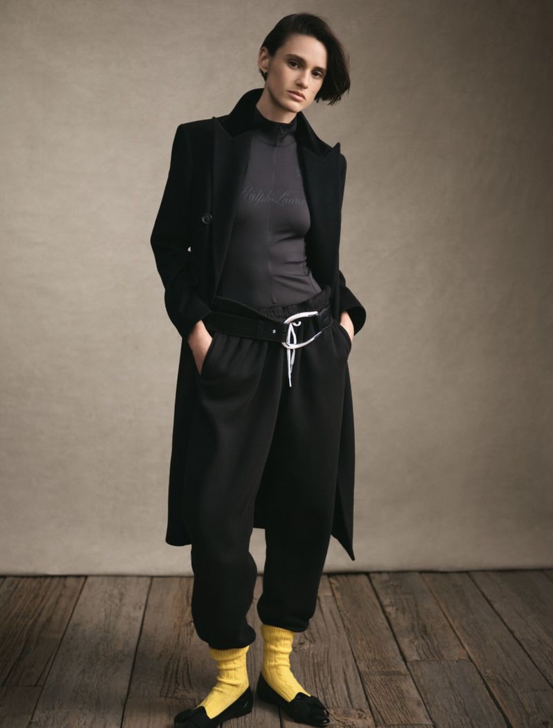Fashion shot of a young adult woman wearing a black coat and matching sweatpants, accessorized with a belt, yellow socks, and black loafers.