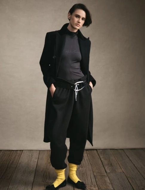 Fashion shot of a young adult woman wearing a black coat and matching sweatpants, accessorized with a belt, yellow socks, and black loafers.