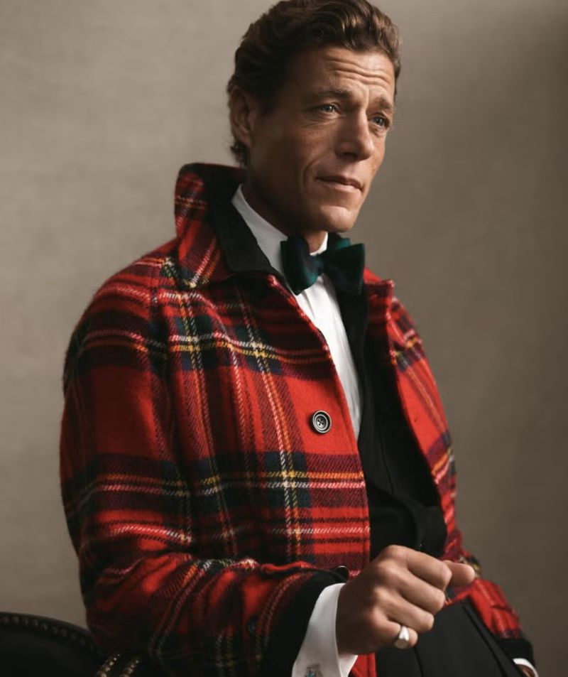 Elegant adult male poses indoors wearing a red plaid jacket with a bow tie and ring, exuding a refined and fashionable appearance.