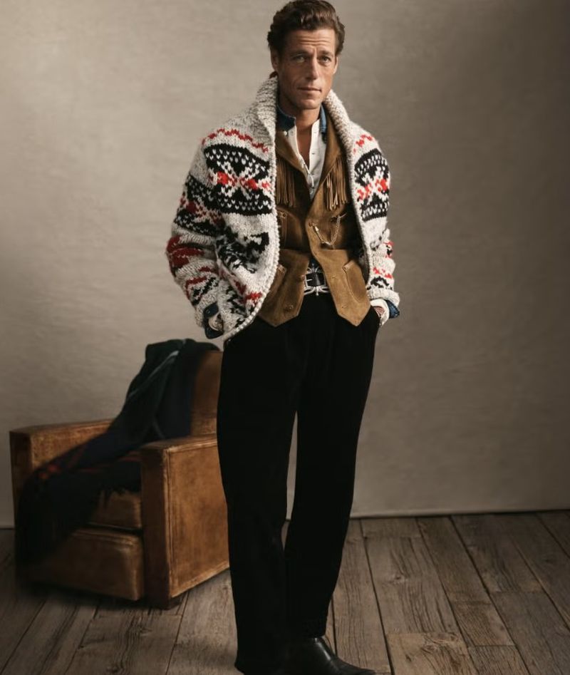 Adult male in fashion shot, wearing pattern sweater, vest, and dress pants, standing on wooden floor with leather chair in background.