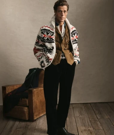 Adult male in fashion shot, wearing pattern sweater, vest, and dress pants, standing on wooden floor with leather chair in background.