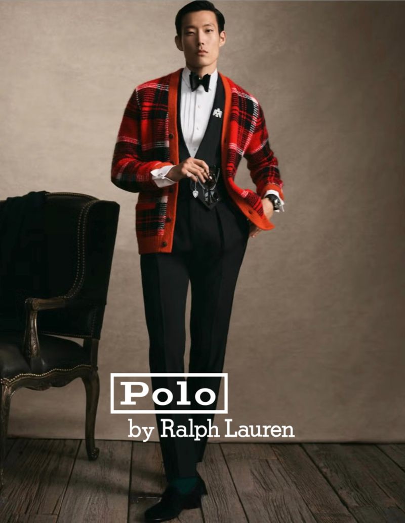 Man in a black tuxedo and a red and black plaid cardigan, standing next to an antique chair, with Polo by Ralph Lauren logo.