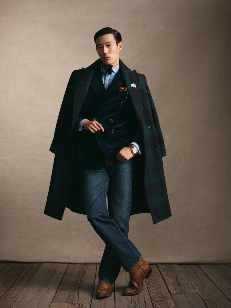 An Asian man in a formal, layered outfit poses in a studio. He wears a plaid coat, velvet suit, blue shirt, jeans and tan boots.