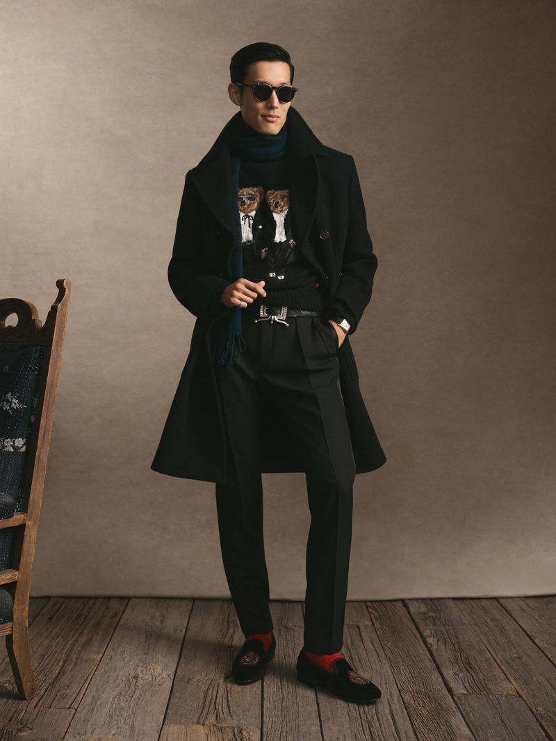 Man wearing a black coat, sunglasses, and sweater standing on a wooden floor in a studio setting