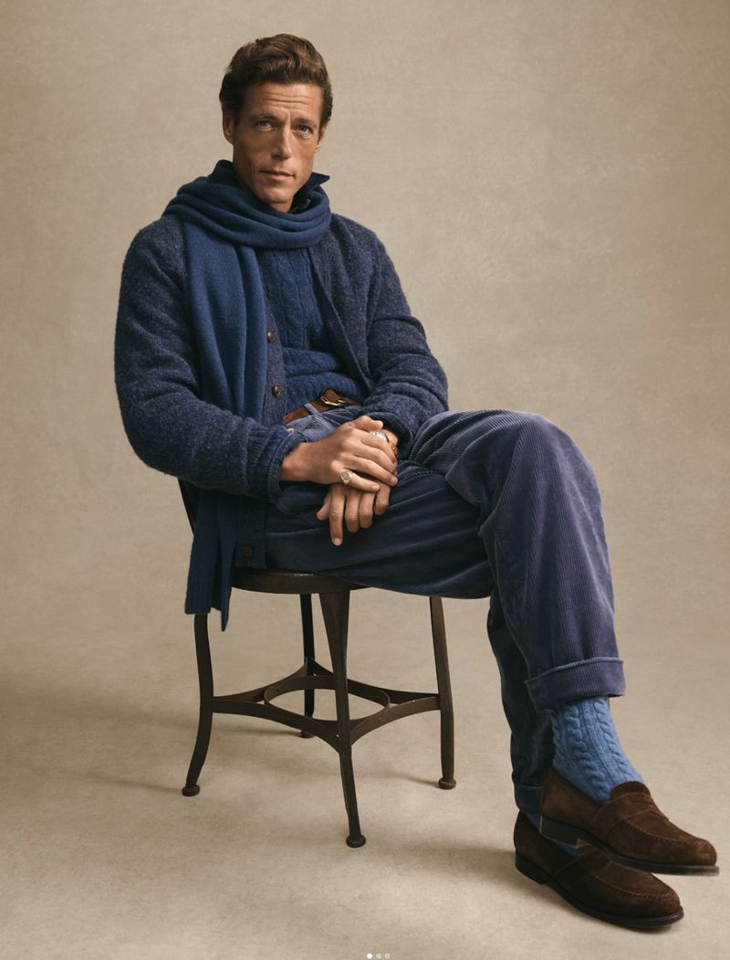 Adult male fashion model poses sitting on stool in a blue corduroy outfit and brown suede loafers against a neutral backdrop in studio lighting.