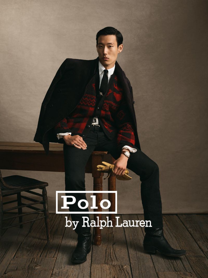A stylish Asian male fashion model poses in black jeans and boots with a red patterned vest under a dark jacket in a studio setting.