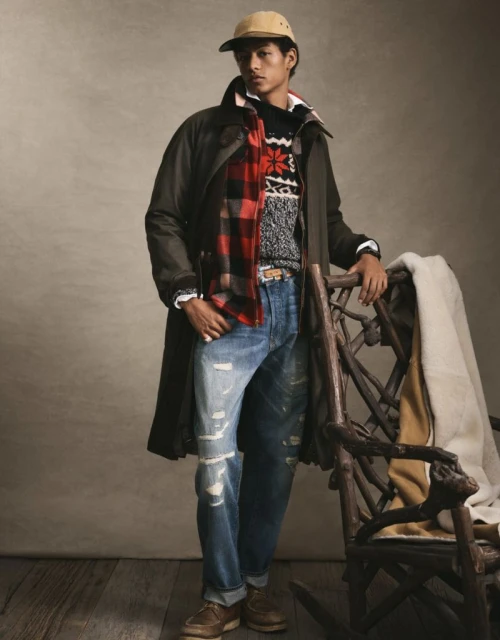 Man in a dark green coat, red plaid scarf, and blue jeans poses near a wooden chair in a warm-toned indoor setting.