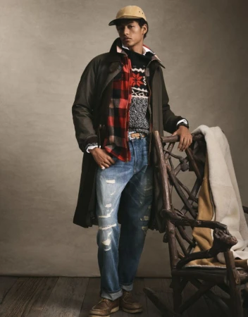 Man in a dark green coat, red plaid scarf, and blue jeans poses near a wooden chair in a warm-toned indoor setting.