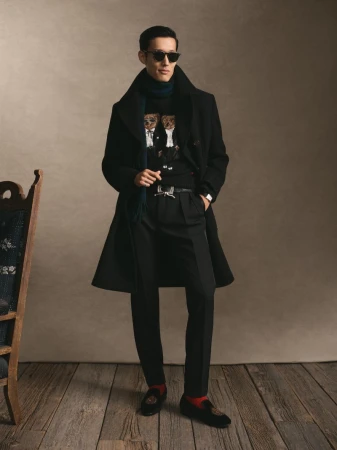 Man wearing a black coat, sunglasses, and sweater standing on a wooden floor in a studio setting