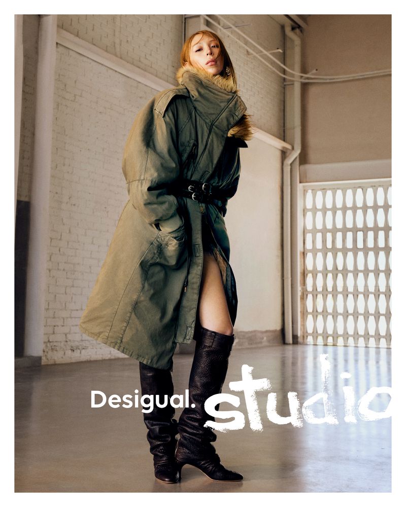 A woman wearing a long green coat and black boots poses in a minimalist studio. The brand name Desigual is displayed in the foreground.