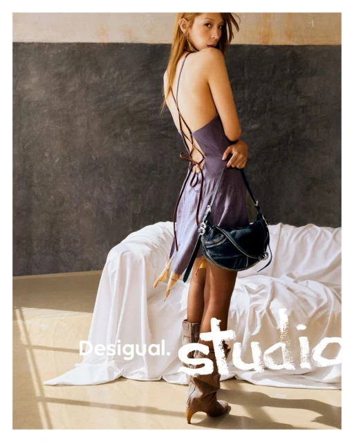 Fashion shot of a woman in a purple dress and boots, carrying a black purse, in front of a neutral backdrop and white couch.