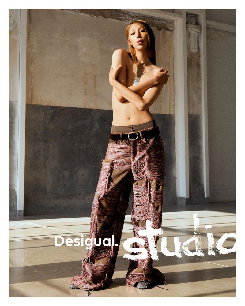 A person poses indoors wearing wide-leg pants with string details, accessorized with a belt and necklace, near a branded graphic.