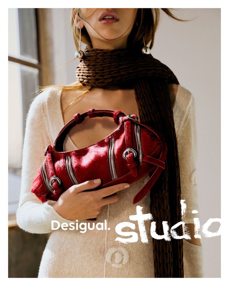 Fashion shot: Model presents a Desigual red patent handbag with zippers, wearing a cardigan, brown scarf, and statement earrings indoors.