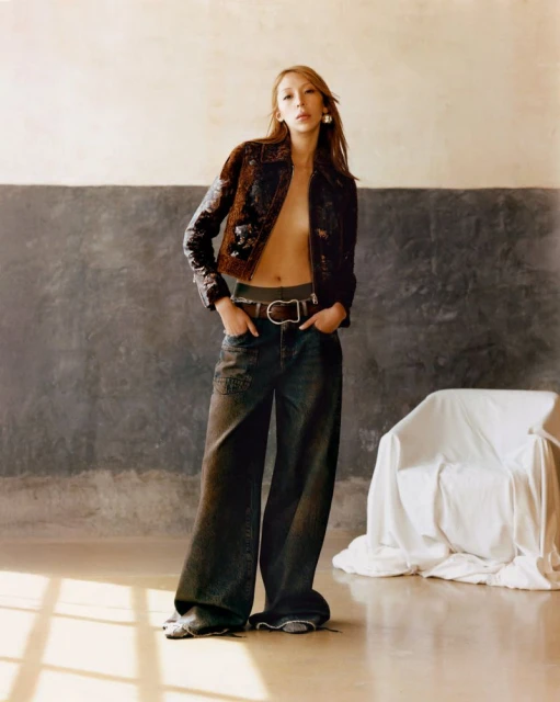 A fashionable woman poses in a brown jacket, dark jeans, and sneakers in a studio setting with a chair covered in white fabric.